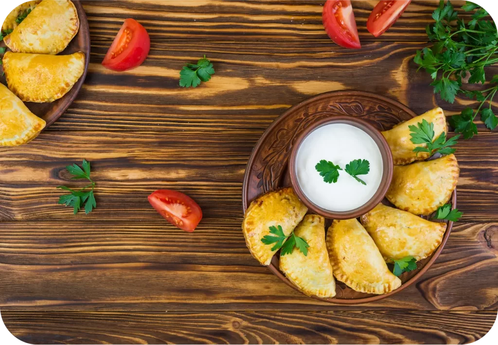 Empanadas doradas al horno servidas con crema agria y decoradas con perejil fresco, acompañadas de tomates en rodajas sobre fondo rústico de madera. Ideal para compartir